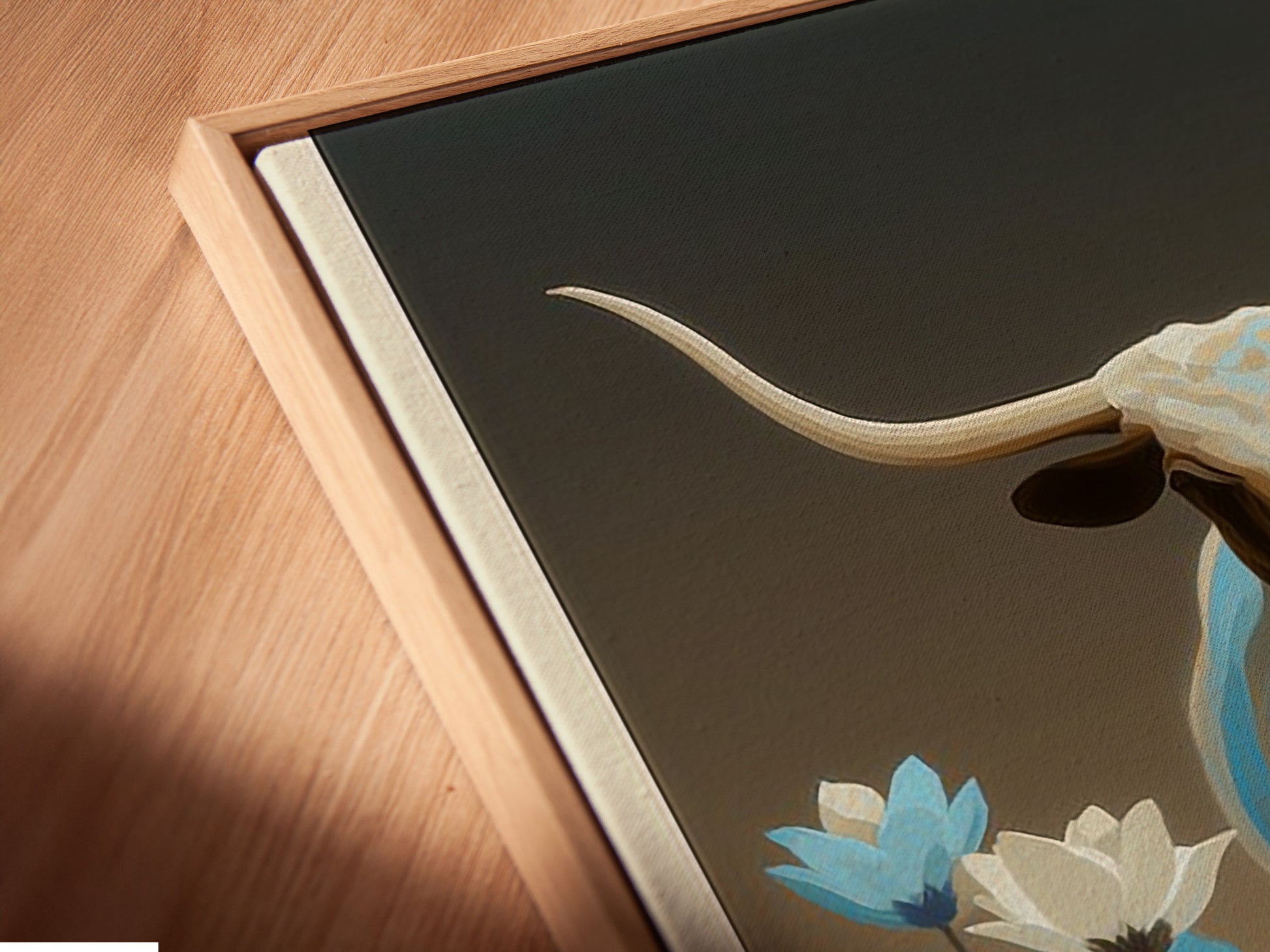 A close-up shot of a canvas print in a floating frame. The longhorn bull artwork features blue wildflowers. Perfect Western wall decor for any room. This bull canvas print is ideal for modern farmhouse decor.