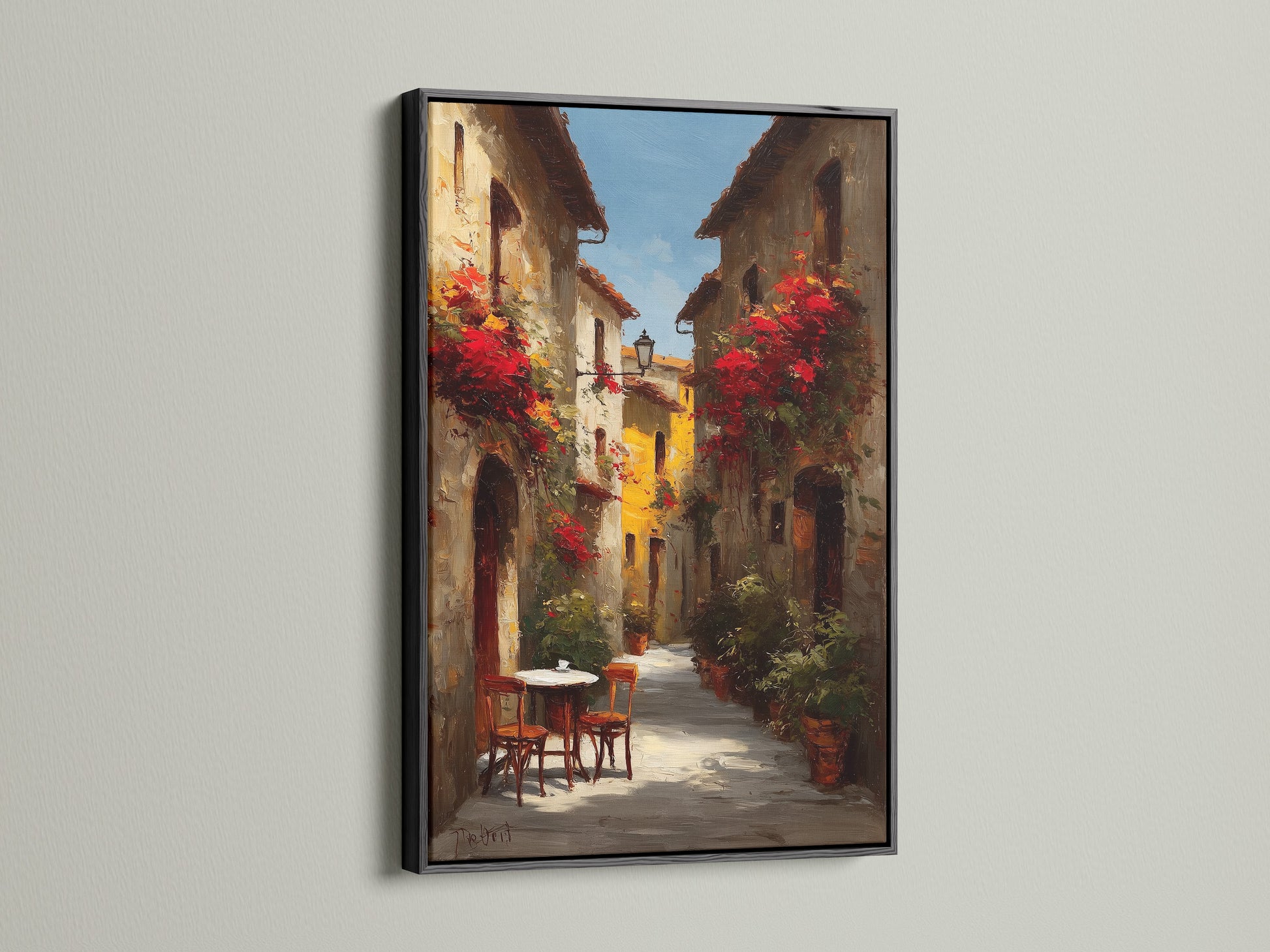 Black frame artwork. The art showcases a lovely Italian street scene with blooming red flowers. A small table and chairs offer a welcoming touch. The artwork is displayed on a white wall. The light accentuates the contrast and details of the art.