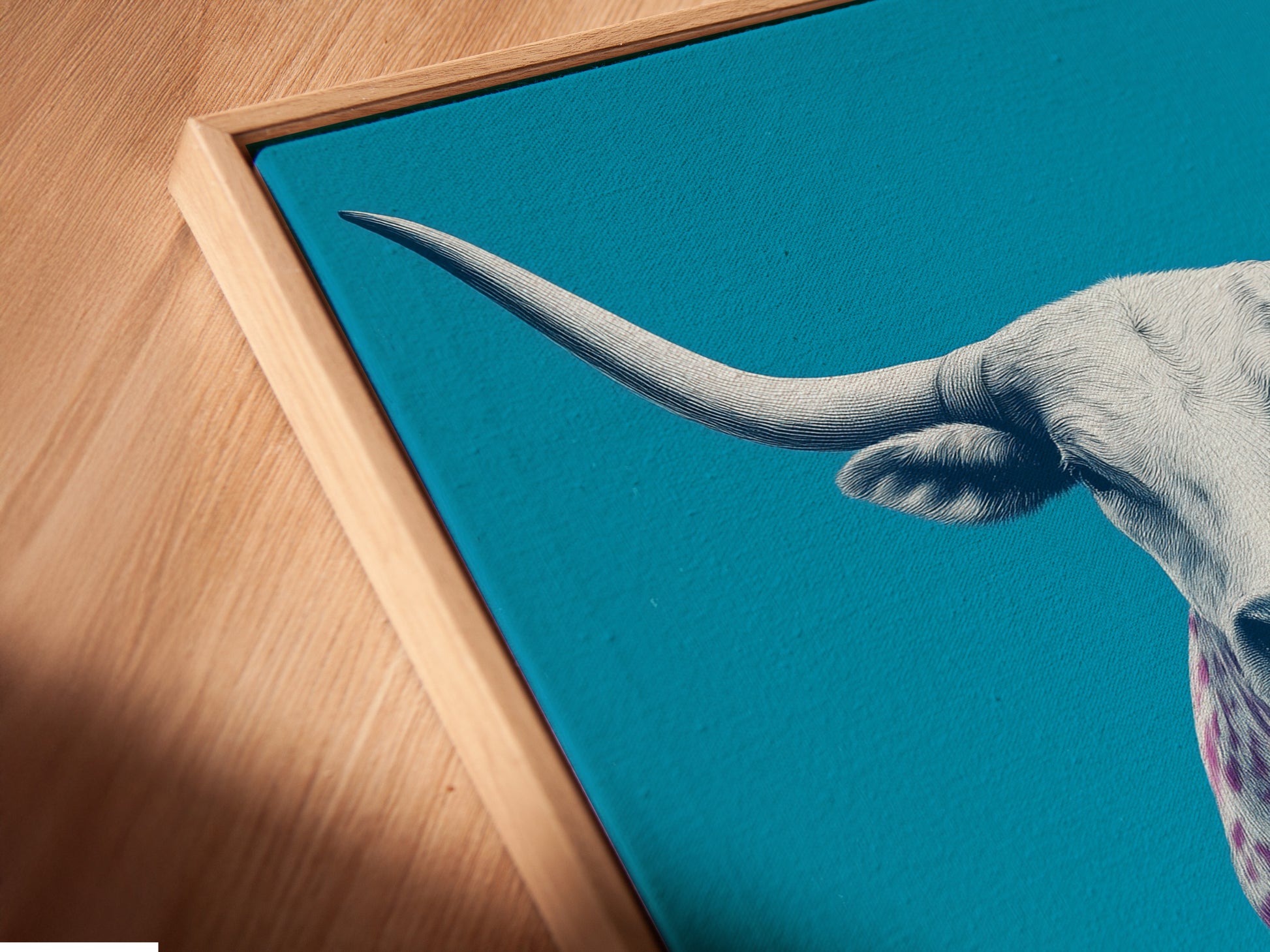 Close-up angled view of a canvas print in a floating frame. The art features a longhorn cow with pink spots on a turquoise background. The floating frame enhances the artwork. It brings a modern touch to the western art piece.