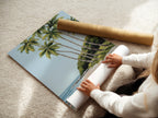 A woman is carefully rolling a fine art poster paper. This poster features a tropical beach scene with palm trees and a boat. This fine art poster paper ensures high quality art. Perfect for adding a coastal touch to your home.