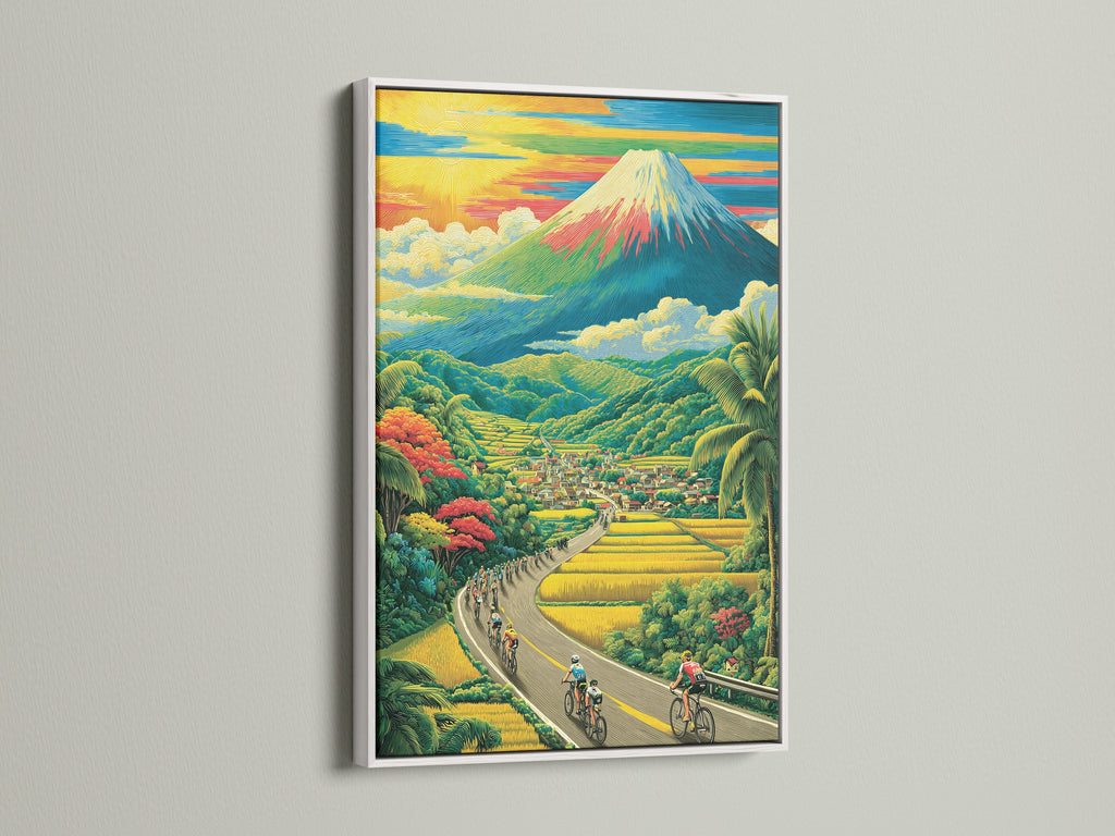 This landscape artwork is showcased in a clean white frame. The artwork features cyclists on a road. A picturesque village and impressive mountain are in the background. The white frame offers a fresh contemporary look.