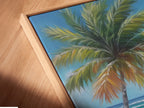 Close-up of a canvas print in a floating frame. The tropical beach artwork features palm tree coastal wall art and a turquoise ocean painting. The artwork captures a soothing coastal vibe perfect for living room wall art. Ready to hang and admire.