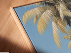 Close up angled view of a canvas print in a floating frame. The art shows palm trees on a white sand beach with a boat in the ocean. This modern framing style adds depth to the tropical beach artwork.