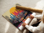 A woman is carefully rolling a fine art poster paper featuring a City Skyline. The artwork captures a sunset over an urban landscape with a drip paint style. This vibrant art print is perfect for adding color to any room. The poster paper ensures high quality.