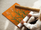 A woman is carefully rolling a fine art poster paper. The paper showcases a floral field artwork. Featuring an orange botanical theme. This modern plant artwork is ready for framing. Perfect for adding a touch of sophistication to any space. Ideal for living rooms bedrooms and offices.