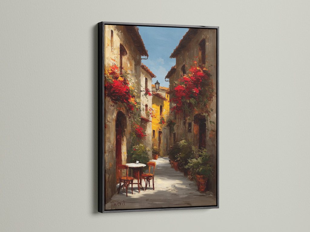 Black frame artwork. The art showcases a lovely Italian street scene with blooming red flowers. A small table and chairs offer a welcoming touch. The artwork is displayed on a white wall. The light accentuates the contrast and details of the art.