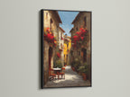 Black frame artwork. The art showcases a lovely Italian street scene with blooming red flowers. A small table and chairs offer a welcoming touch. The artwork is displayed on a white wall. The light accentuates the contrast and details of the art.