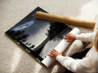 A woman is rolling a fine art poster paper. The paper features a Night Sky Landscape. This allows for easy and safe shipping of the galaxy art. The lake wall art is ready to be framed. High-quality celestial art for your home.