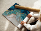 A woman carefully rolls a fine art poster paper. The paper features a captivating blue abstract artwork. The fluid acrylic texture and gold accents add a touch of luxury. This poster is perfect for modern bedroom decor. Ideal for those seeking elevated contemporary wall decor.