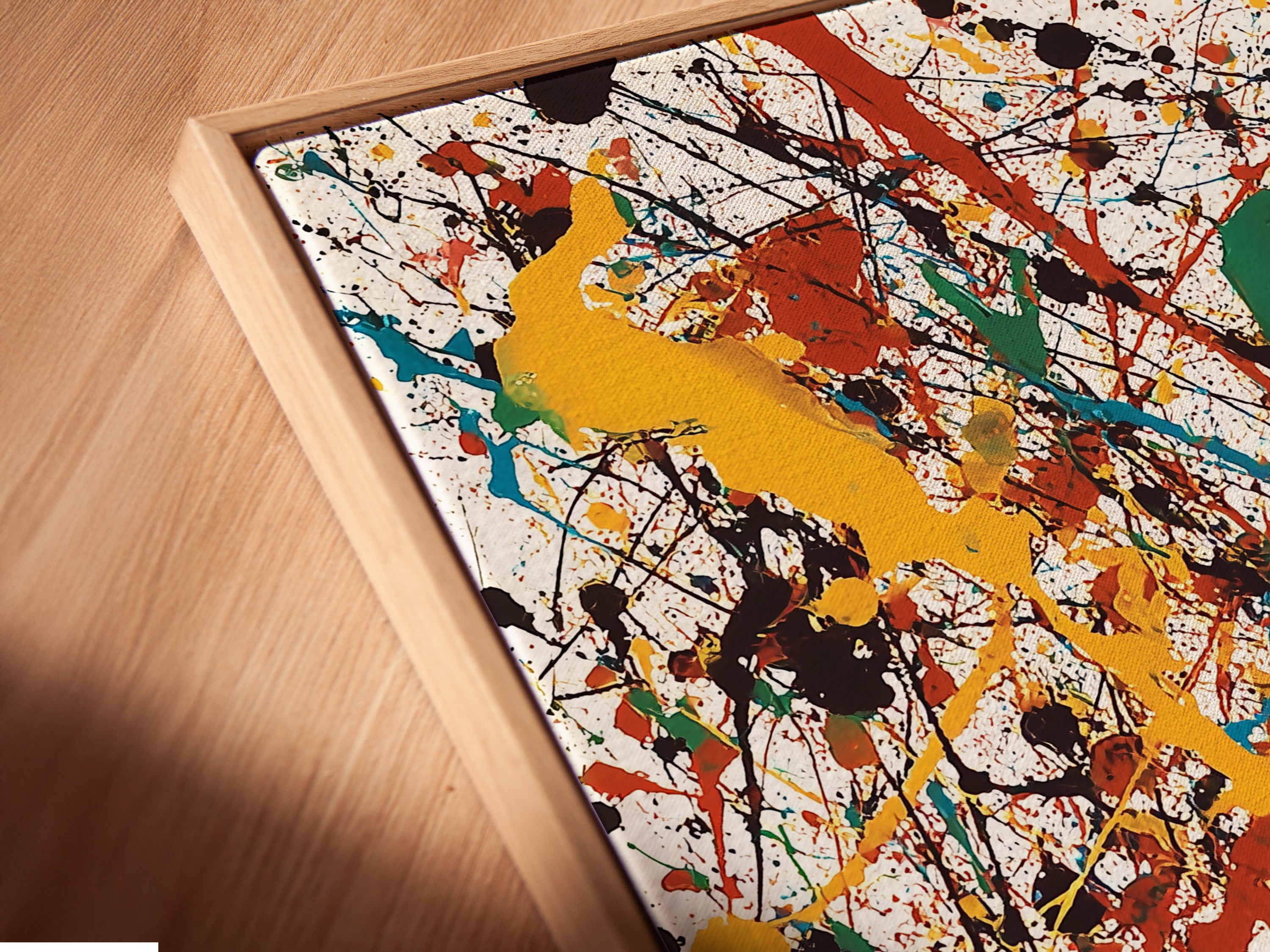 A close-up angled view of a canvas print in a floating frame. The frame creates a unique visual effect. The artwork features splatters of yellow teal red green and black paint. The floating frame makes the art stand out.