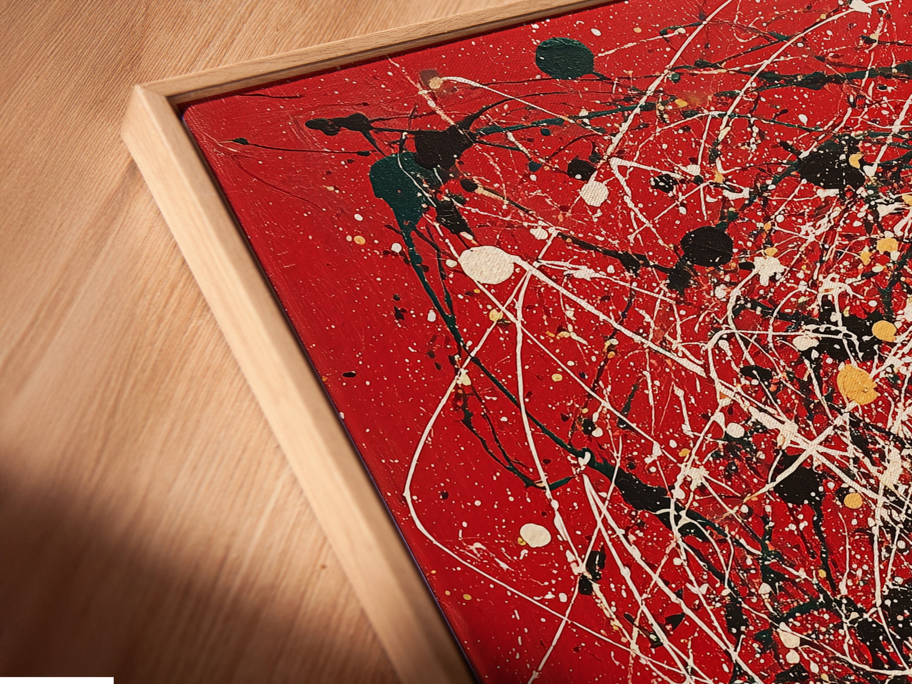 A close-up angled view of a canvas print in a floating frame. The abstract artwork features red canvas with splatter art. Paint drips add a modern and contemporary touch. This piece enhances any home decor. Ideal for those seeking unique and stylish wall art. Perfect for living rooms and offices.