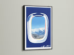 Artwork featuring an airplane window view in a black frame. The print offers a scenic landscape with snow-capped mountains and a blue sky. This modern travel art is a wanderlust decor piece. A unique aviation art piece.