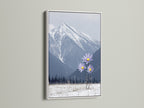 A mountain flower canvas print is elegantly displayed in a white frame. This winter print captures a stunning landscape. Snowy mountains and lilac flowers create a serene bedroom art piece. The modern decor adds a fresh touch to your space.
