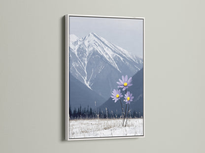 A mountain flower canvas print is elegantly displayed in a white frame. This winter print captures a stunning landscape. Snowy mountains and lilac flowers create a serene bedroom art piece. The modern decor adds a fresh touch to your space.