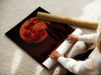 A woman is carefully rolling a fine art poster paper. The paper features a captivating red moon and forest landscape. Perfect for creating high-quality celestial art prints. Order yours today and make some art.