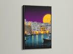 The black frame provides a striking contrast to the coastal village artwork. The artwork portrays a vibrant Mediterranean harbor scene. The harbor is filled with boats and illuminated buildings. It is a stunning piece of evening wall art.