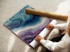 A woman is carefully rolling a fine art poster paper. The paper features an Abstract Ocean Wave artwork. It displays beautiful teal and purple fluid art. This poster is perfect for contemporary coastal decor. It provides a modern abstract touch to any living space. Explore our fine art prints today