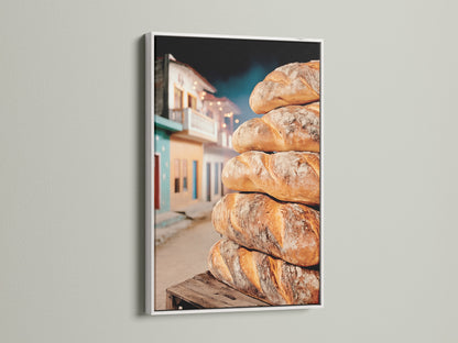 A stack of bread against a rustic street scene is featured in a white frame. This kitchen artwork adds a touch of elegance. Enhance your cafe decor with food photography. It brings culinary charm.