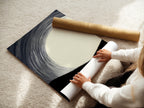 A woman carefully rolls a fine art poster paper. The paper features a minimalist artwork. The artwork is an abstract landscape in beige black and white. High-quality materials ensure a stunning piece. Perfect for adding a touch of elegance to any interior.