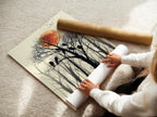 A woman is carefully rolling a fine art poster paper print. The artwork features birds on a tree with a red sun and forest silhouette. This nature wall art is a great gift for bird watchers.