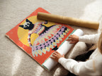 A woman is carefully rolling a fine art poster paper featuring whimsical cat art. The print is vibrant with orange and yellow hues. Perfect for animal wall art or quirky cat decor. This folk art is ideal for any room.
