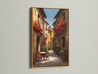 Gold frame artwork. The art presents a delightful Italian street scene decorated with red flowers. A cozy table and chairs invite you in. The artwork is displayed on a white wall. The lighting highlights the elegant gold frame and the art's beauty.