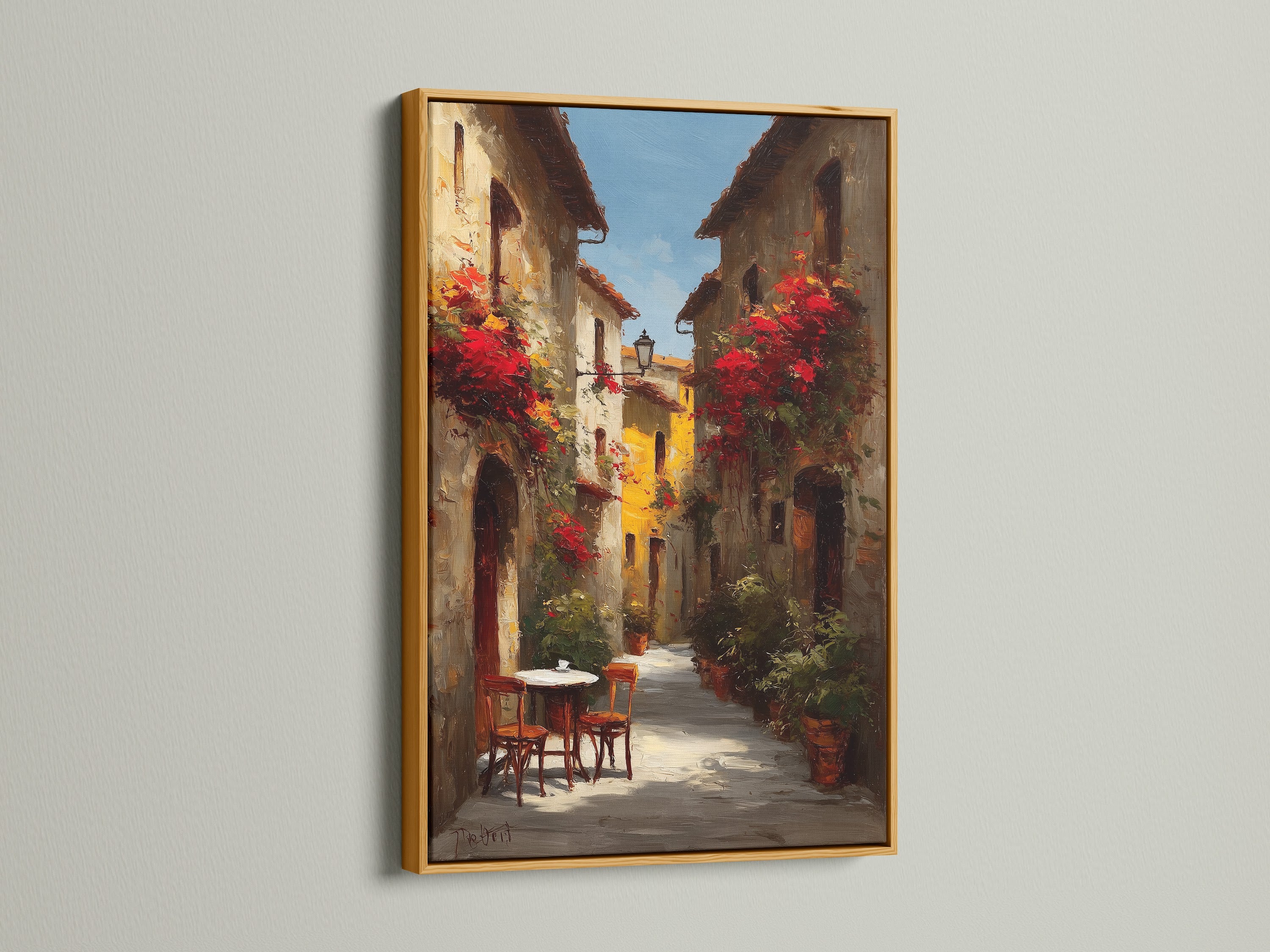 Gold frame artwork. The art presents a delightful Italian street scene decorated with red flowers. A cozy table and chairs invite you in. The artwork is displayed on a white wall. The lighting highlights the elegant gold frame and the art's beauty.