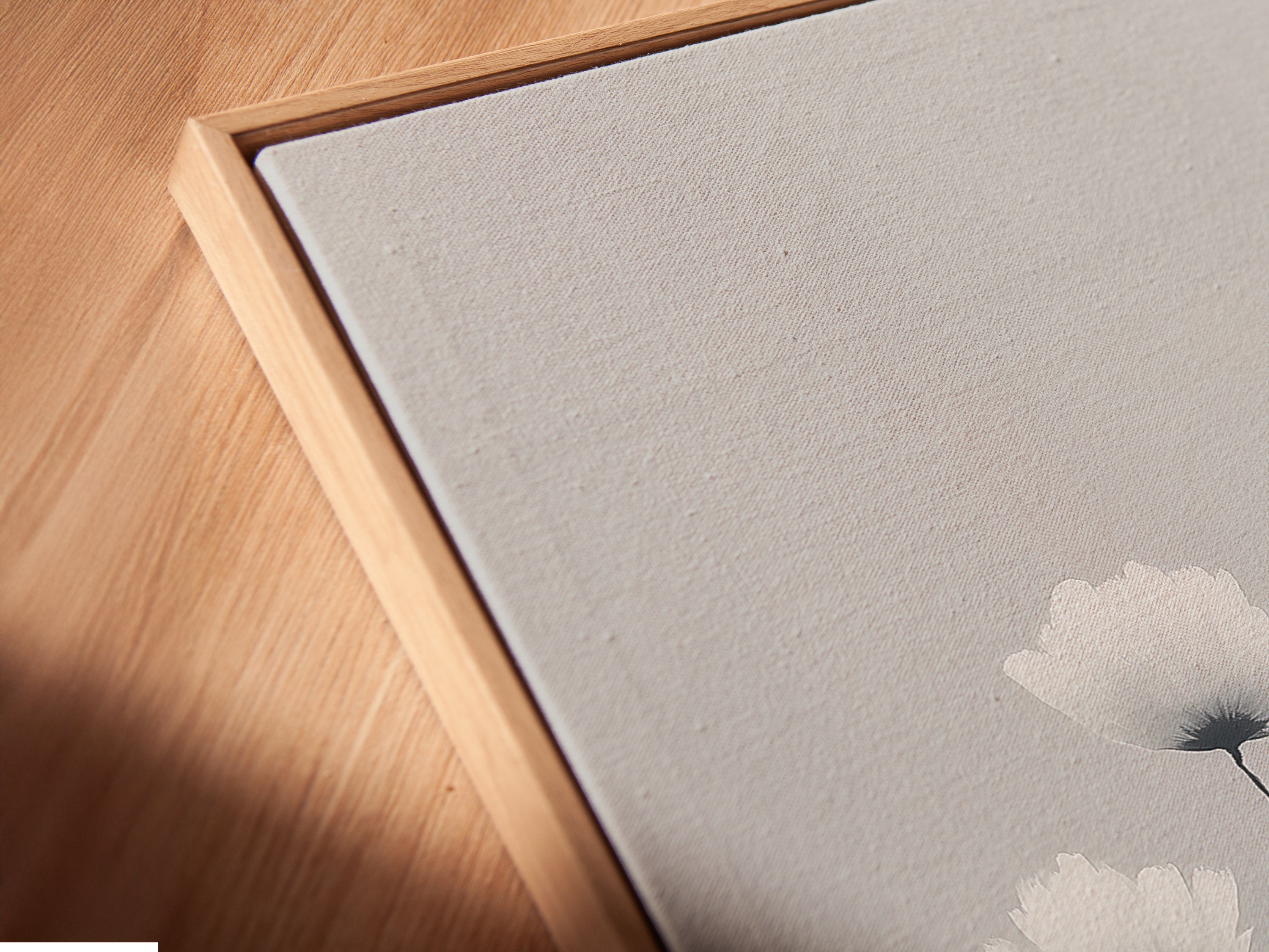 A close-up shows a canvas print in a floating frame. The minimalist flower artwork features soft gray and white tones. This modern canvas art adds depth to any room. Floating frames give the botanical print a gallery-like appearance elevating the artwork.