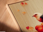 Close-up of a Bird and Apples canvas print in a floating frame. The artwork is a rustic fall still life. This piece is ideal for kitchen decor. The floating frame gives the art a modern look. It showcases a bird perched on a bucket of red apples. This close-up highlights the artwork's fine details.
