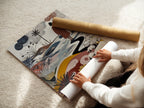 A woman is carefully rolling a fine art poster paper. The poster paper is of high quality ensuring vivid colors. The artwork features a figure walking along a winding path. Sheep graze in a field of flowers. Order your fine art poster today.