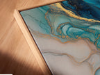 A close-up angled view reveals the intricate details of a canvas print in a floating frame. The blue abstract art features fluid acrylic textures and gold accents. This piece adds a modern coastal touch to any living space. The artwork enhances modern bedroom decor with its teal and marble print.