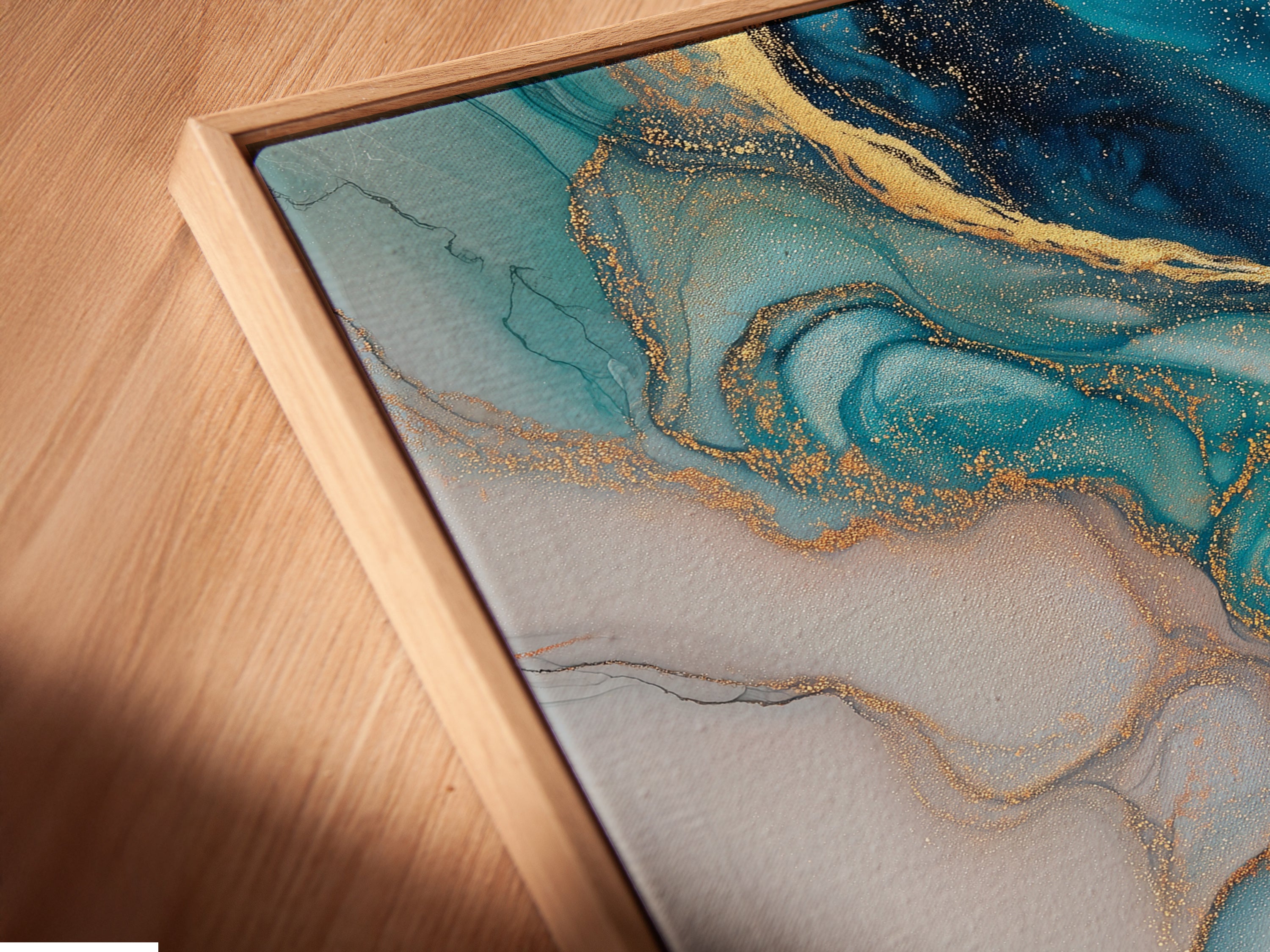 A close-up angled view reveals the intricate details of a canvas print in a floating frame. The blue abstract art features fluid acrylic textures and gold accents. This piece adds a modern coastal touch to any living space. The artwork enhances modern bedroom decor with its teal and marble print.