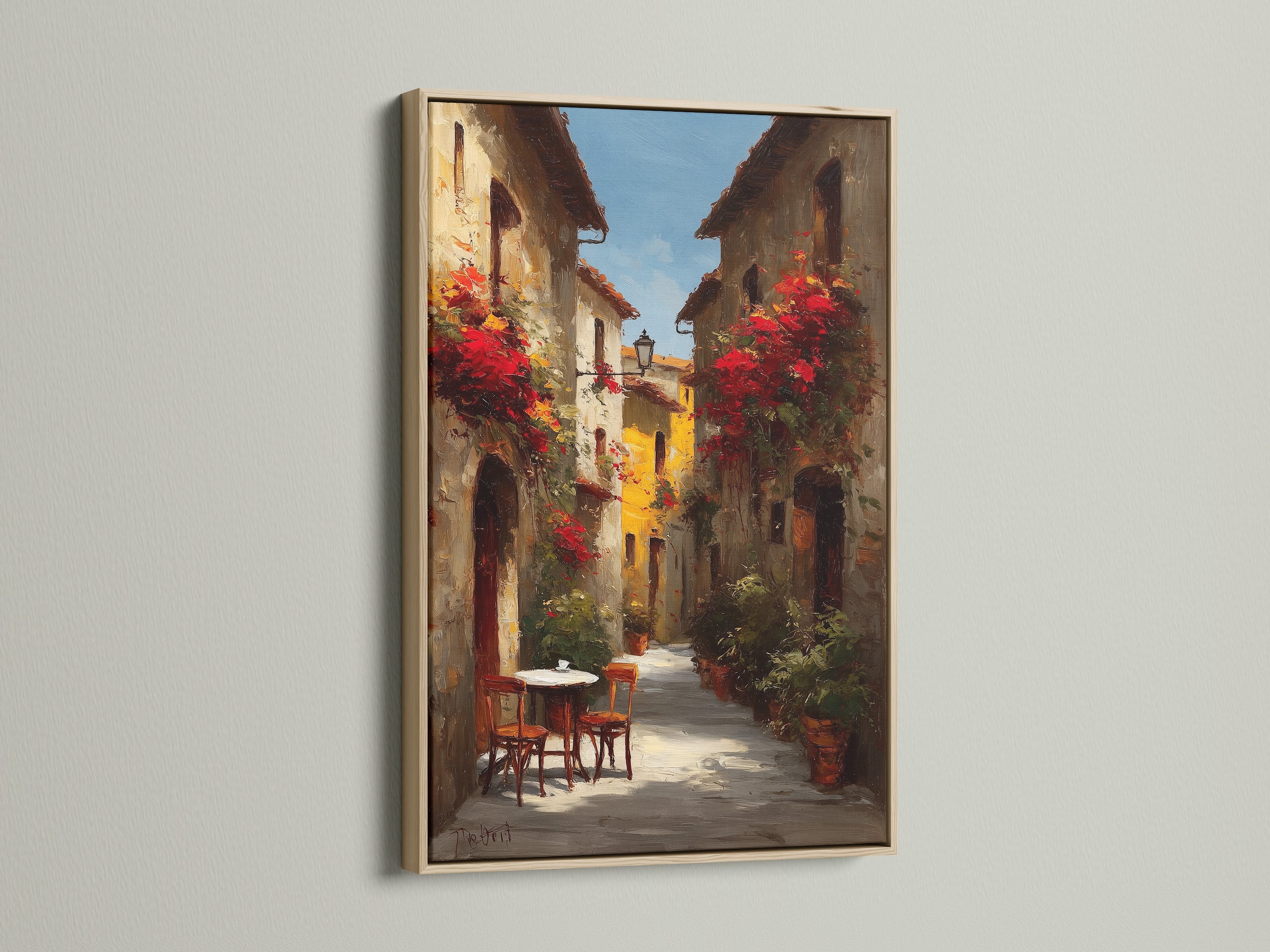 Oak frame artwork. The art captures a picturesque Italian street filled with vibrant red flowers. A small table and chairs create a cozy atmosphere. The artwork is displayed on a white wall. The light emphasizes the details of the art and frame.
