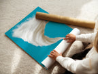 A woman carefully rolls a fine art poster paper showcasing the surreal coffee cup storm artwork. This turquoise sky and beach art print is perfect for modern home decor. Ideal for art collectors and coffee lovers. Add an imaginative touch to your living space with this unique artwork.