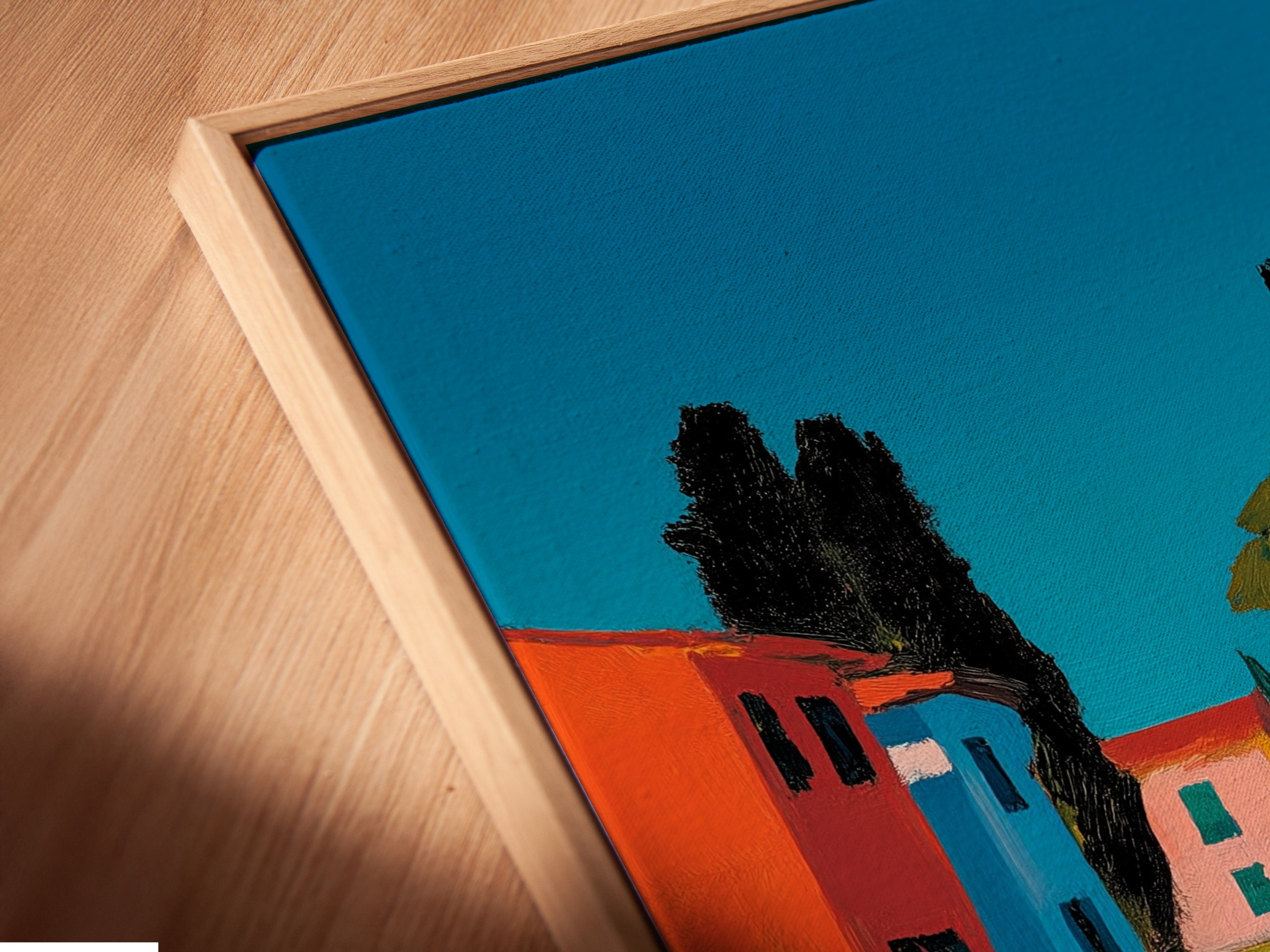 Angled close up of a canvas print in a floating frame. The art features a path leading to colorful buildings. It is a coastal scene under a bright sky. The floating frame gives a modern gallery look. It highlights the canvas texture.