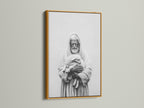 A framed artwork hangs on a light beige wall. The artwork has a gold frame. The frame is thin and modern. The art print is a black and white image of a man holding a lamb. Purchase this beautiful artwork today.