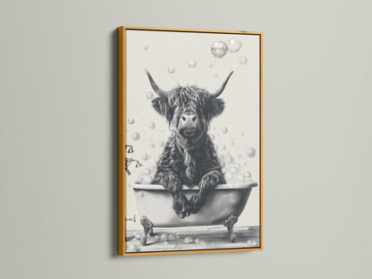 A framed Highland Cow art print is displayed on a white wall. The artwork features a black and white image of a cow relaxing in a bathtub filled with bubbles. The frame is a rich gold color. It complements Country Farmhouse style bathroom art or wall decor.