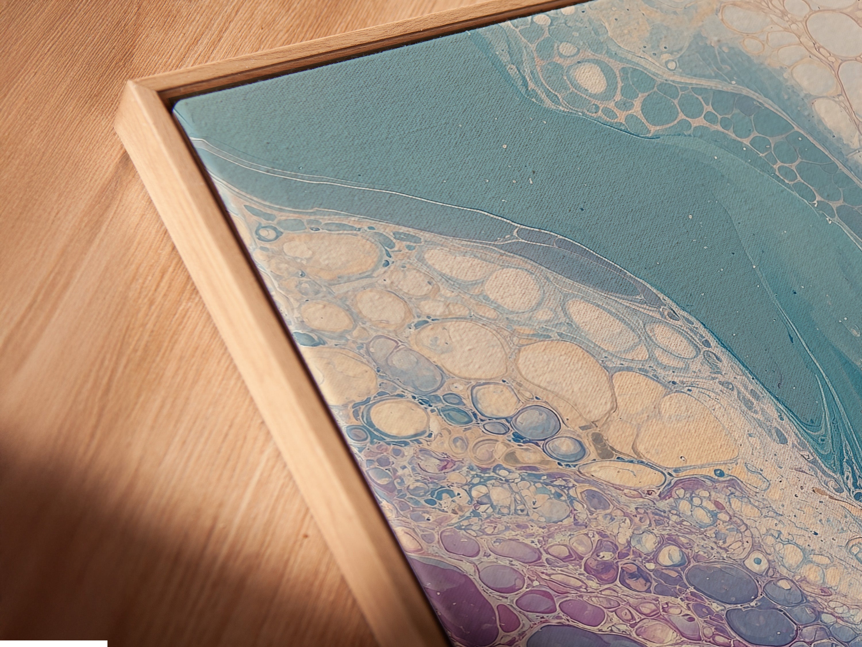 A close-up angled view of a canvas print in a floating frame is shown. The abstract fluid artwork features teal and purple tones. It creates a mesmerizing marble effect. This contemporary piece is perfect for modern home interiors. Elevate your space with this unique art.