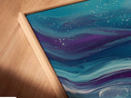 A close-up angled view of a canvas print in a floating frame. The artwork shows teal and purple fluid art. Great coastal wall art for any bedroom decor. It's modern beach artwork.